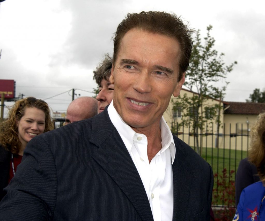 Arnold Schwarzeneggar steht im schwarzen Anzug vor einem Podium.