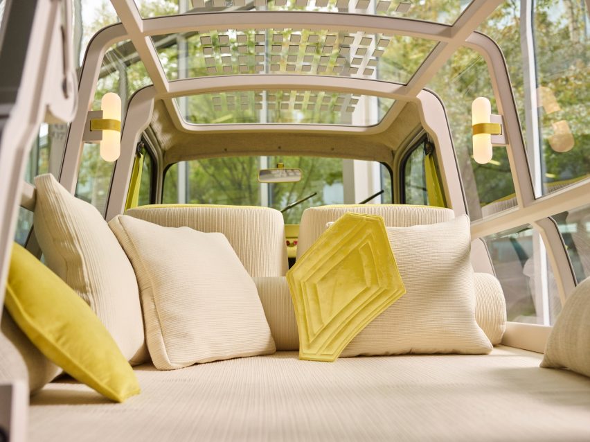 Bett im Kofferraum eines Autos mit cremefarbener Chenille-Polsterung, Kissen und umlaufenden transparenten Paneelen mit Blick auf einen Wald