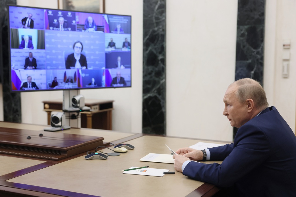 Putin sitzt an einem Schreibtisch und sieht sich einer im Fernsehen übertragenen Videokonferenz gegenüber.